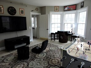 A living room with a television, a futon, a patterned rug, a futsal court, and a door leading to another room. - Cabin Retreat for Nature and City Exploration near Sebago Lake State Park, ME (Casco)