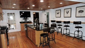 Una cocina con suelo de madera, una isla central con taburetes, un horno empotrado y un televisor en la pared.