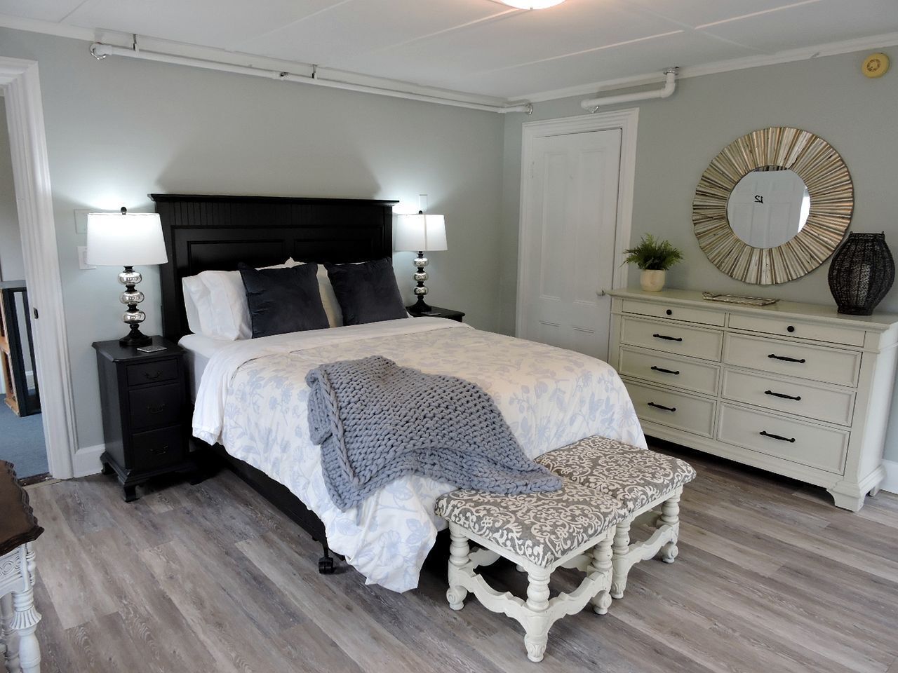 Um quarto com cama, mesas de cabeceira, um banquinho, cômoda e um espelho.