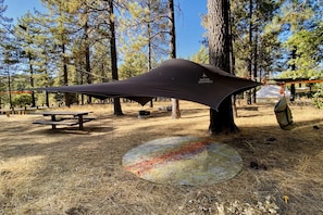 A camping area with a tarp shelter, picnic tables, and a fire pit.