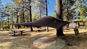 Un área de camping con un refugio de lona, mesas de picnic y una hoguera.