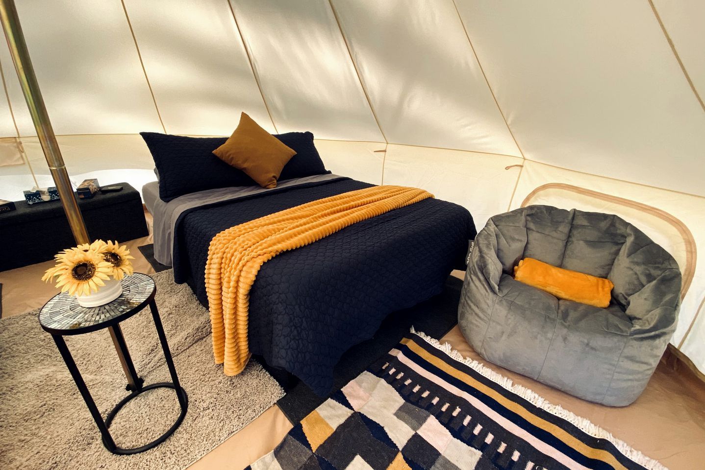 Een tentkamer met een bed, een zakstoel, een klein tafeltje met een vaas zonnebloemen en een kamerkleed met dessin.