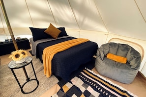 Una habitación tipo tienda de campaña con una cama, un sillón de mimbre, una pequeña mesa con un jarrón de girasoles y una alfombra estampada.