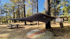 A camping area with a tarp shelter, picnic tables, and a fire pit.