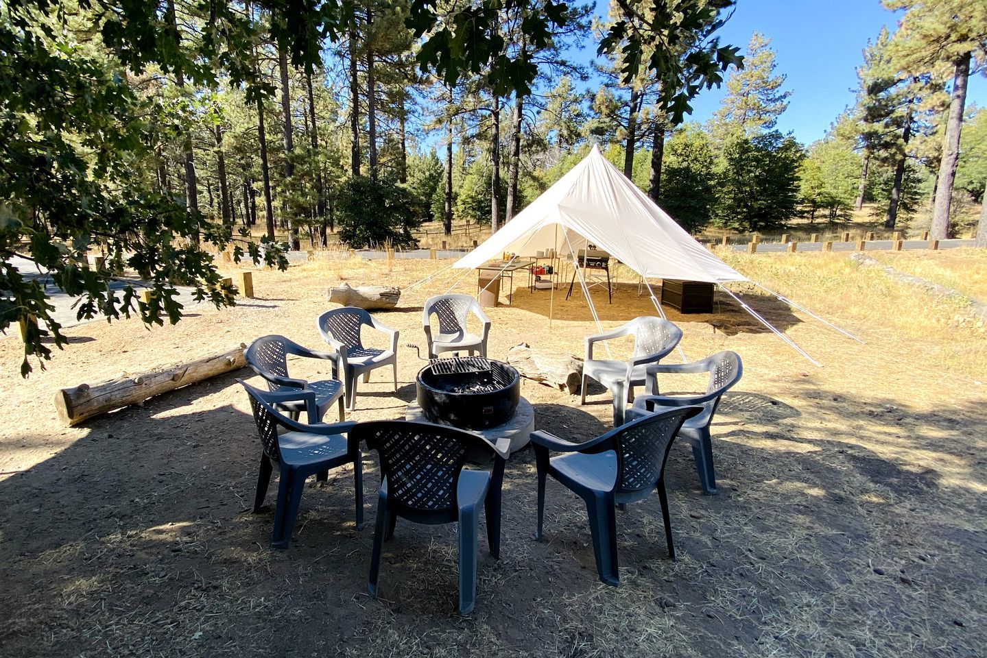 Een camping met een tent, een vuurkuil en plastic stoelen rondom een tafel.