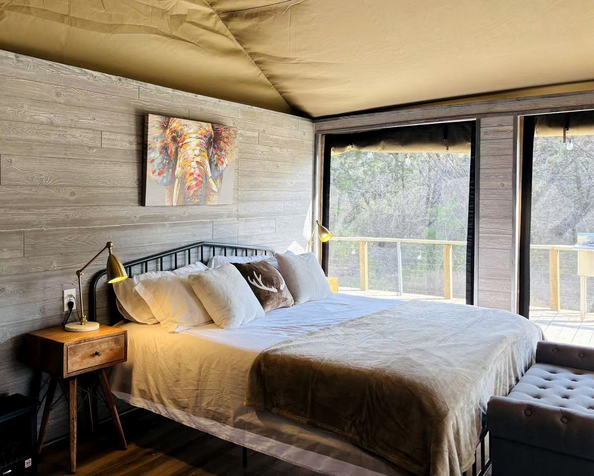 Een slaapkamer met een groot bed, een nachtkastje met een lamp, een schilderij aan de muur en een raam met uitzicht op bomen.