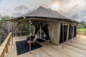 A tented outdoor area with a wooden deck, a table, chairs, and a view of the surrounding trees.