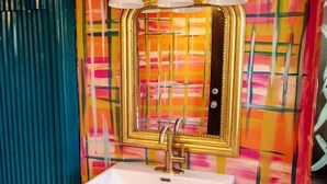 Een badkamer met een witte wastafel, gouden details en een kleurrijk abstract muurontwerp.