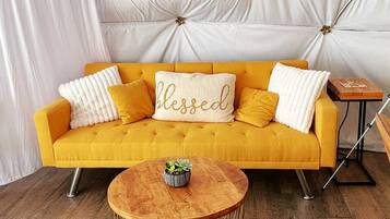 A mustard yellow sofa with a 'blessed' pillow, a small wooden side table, and a round coffee table with a plant.