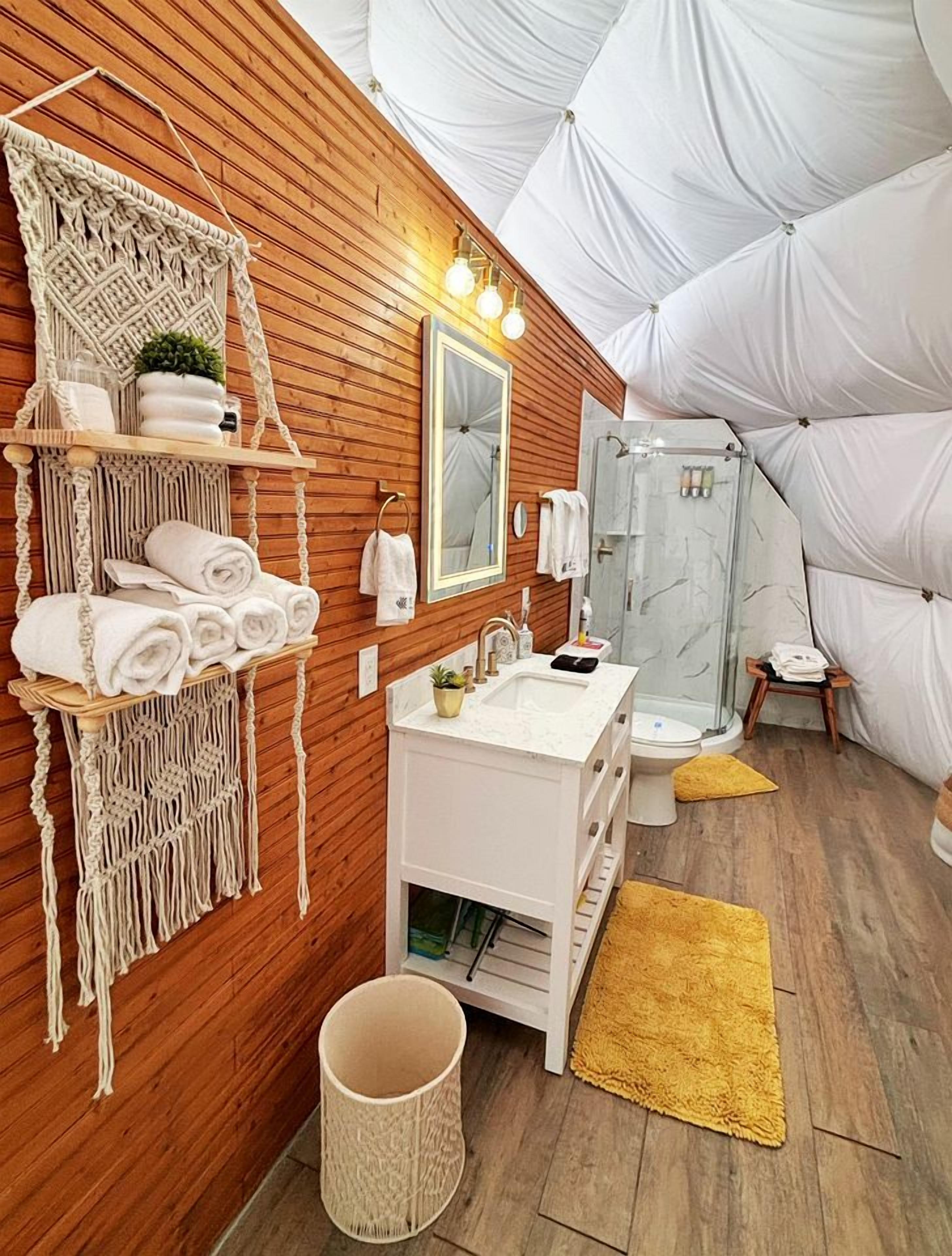 Ein modernes Badezimmer mit Holzvertäfelung, einem weißen Waschtisch, einem großen Spiegel, einer Dusche und einem Wandbehang aus Makramee.