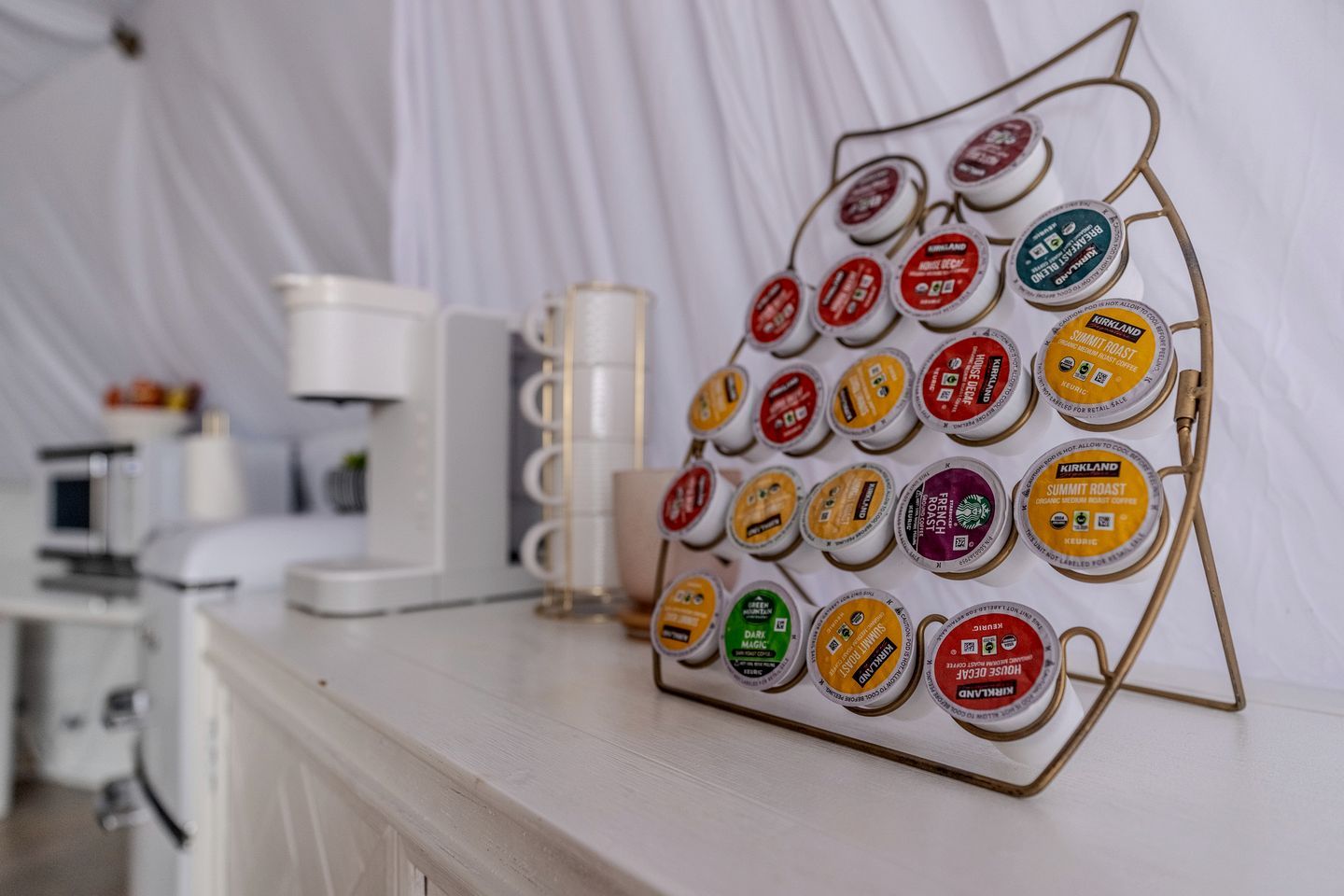 A coffee station with a metal rack holding various coffee pods, a white coffee machine, and a white countertop.