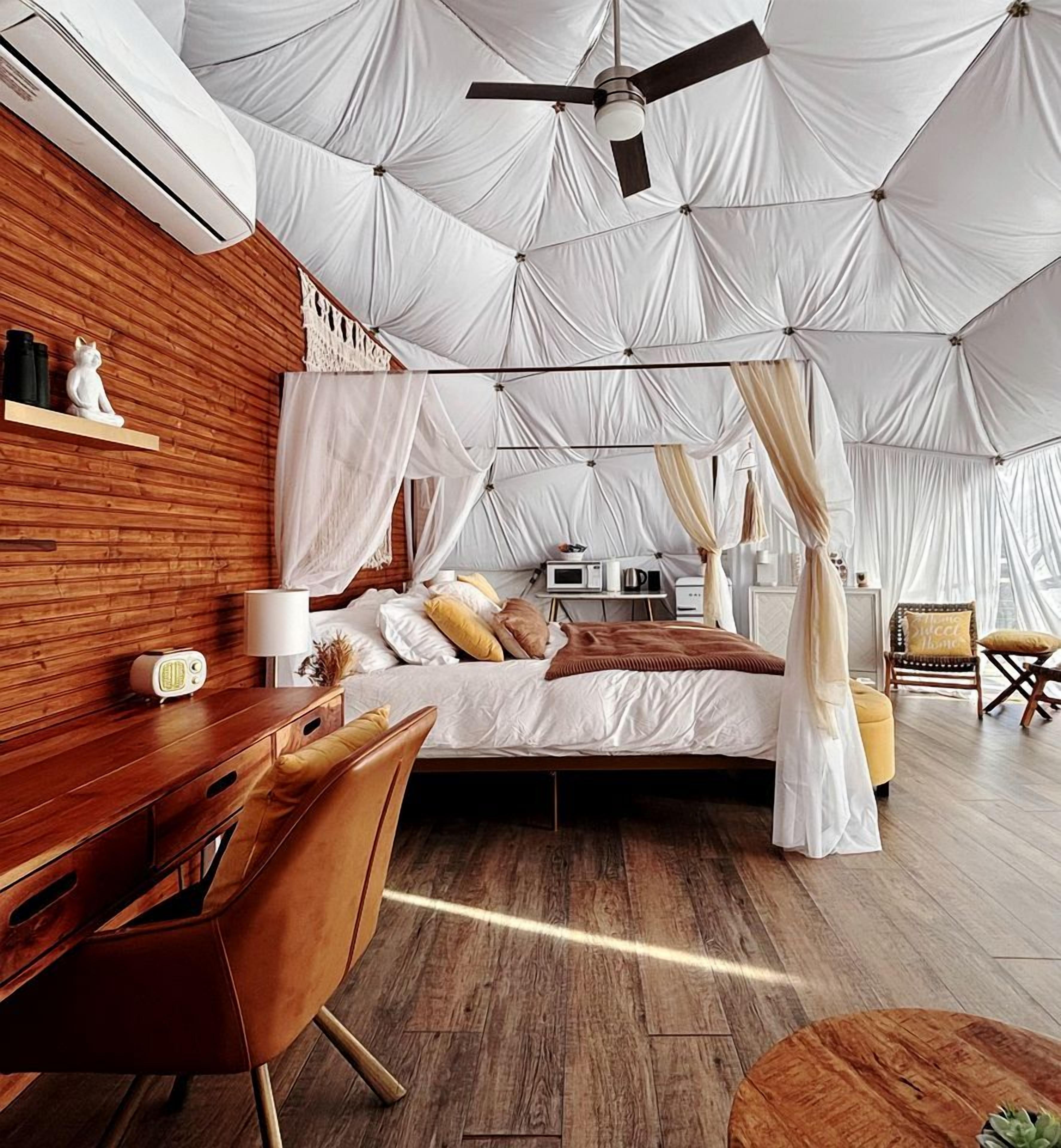 A bedroom with a canopy bed, wooden flooring, a desk with a chair, and a bedside table with a lamp.
