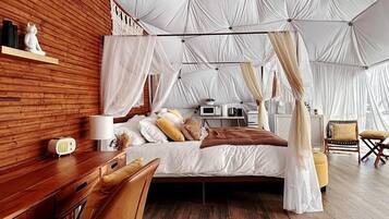 A bedroom with a canopy bed, wooden flooring, a desk with a chair, and a bedside table with a lamp.