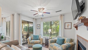 A spacious living room with a ceiling fan, large windows, a fireplace, and a television mounted on the wall. - Charming 1-Bedroom Condo at West Hyde Park – Cozy & Convenient! (Myrtle Beach)