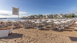 Ein Strand mit Liegestühlen und Sonnenschirmen, eine Flagge und ein Steg im Hintergrund.
