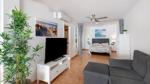 Ein modernes Wohnzimmer mit Flachbildfernseher, einem grauen Sofa, einer großen Bambuspflanze und ein Schlafzimmer mit Bett und Deckenventilator.