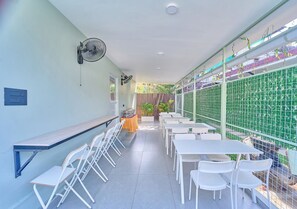 A corridor with white folding chairs and tables, a fan on the wall, and a green fence with plants. - Kayal Cove Resort by VOYE HOMES (Alappuzha)