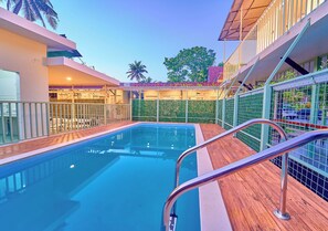 A swimming pool with a wooden deck and metal railings, surrounded by a building with balconies and a green fence. - Kayal Cove Resort by VOYE HOMES (Alappuzha)