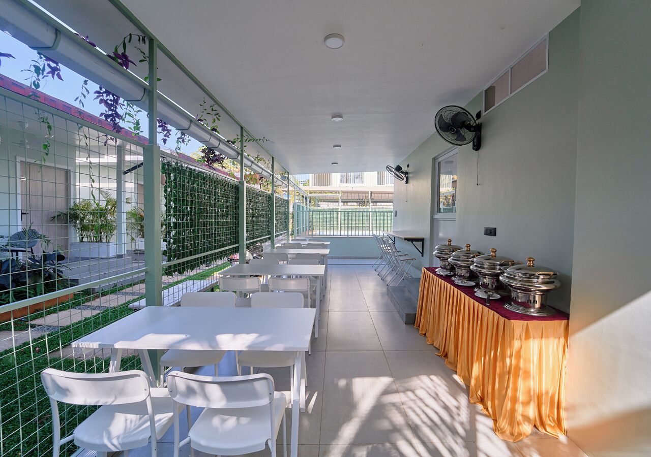 A covered outdoor dining area with tables and chairs, a buffet line with food containers, and a view of the street through a green fence.