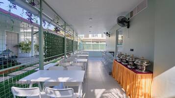 A covered outdoor dining area with tables and chairs, a buffet line with food containers, and a view of the street through a green fence.