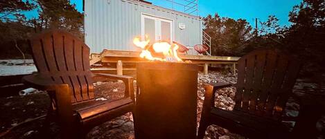 A metal building with a door, a fire pit with flames, and two Adirondack chairs.