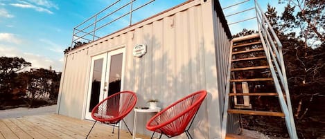 A small, modern cabin with a wooden deck, two red chairs, a table, and a staircase leading to a loft area.
