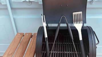 A portable grill with a lid open, two barbecue utensils, and a wooden tray.
