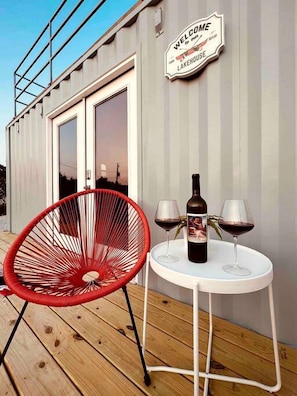 Een terras met een rode stoel, een tafel met twee wijnglazen en een fles, en een bord met de tekst 'Welcome to our Lakehouse'.