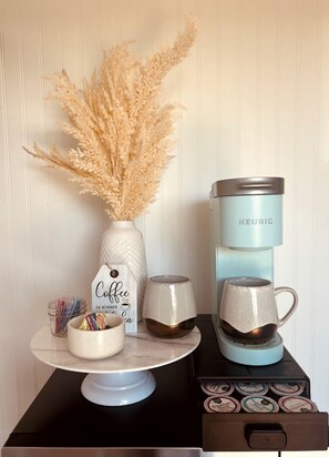 Una cafetera, un florero con plantas secas, una taza de café y una jarra sobre una mesa.