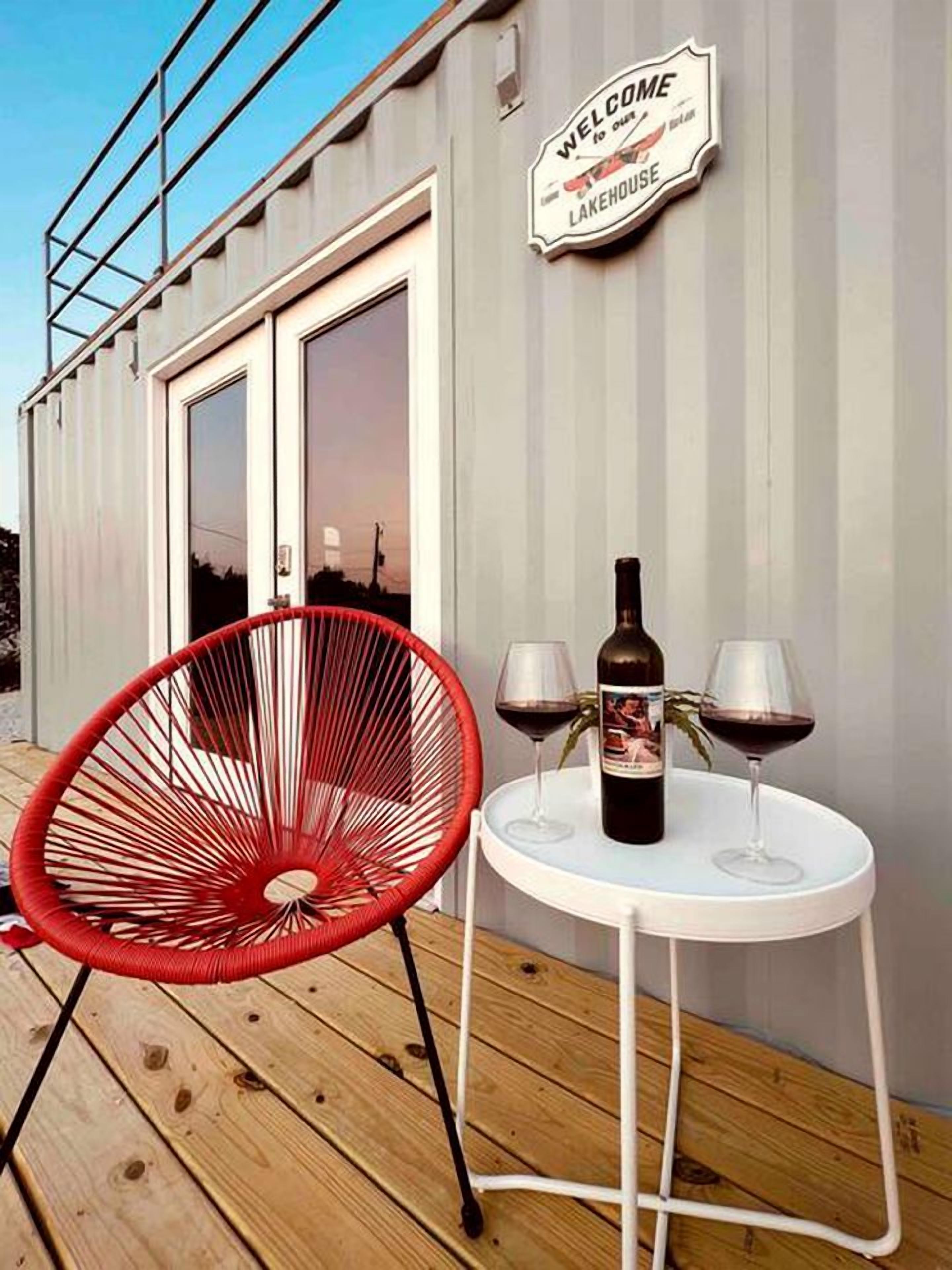 Une terrasse avec une chaise rouge, une table avec deux verres à vin et une bouteille, et une pancarte indiquant : « Bienvenue dans notre maison au bord du lac ».