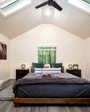 A bedroom with a sloped ceiling, a bed with a wooden frame, two bedside tables, a window with a view of greenery, and a ceiling fan.