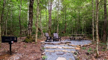 Un área boscosa con un sendero de grava, dos sillas tipo Adirondack, una mesa de picnic y un buzón.