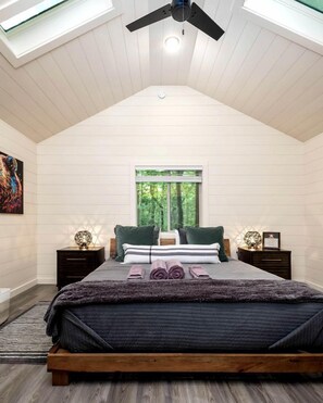 A bedroom with a sloped ceiling, a bed with a wooden frame, two bedside tables, a window with a view of greenery, and a ceiling fan.