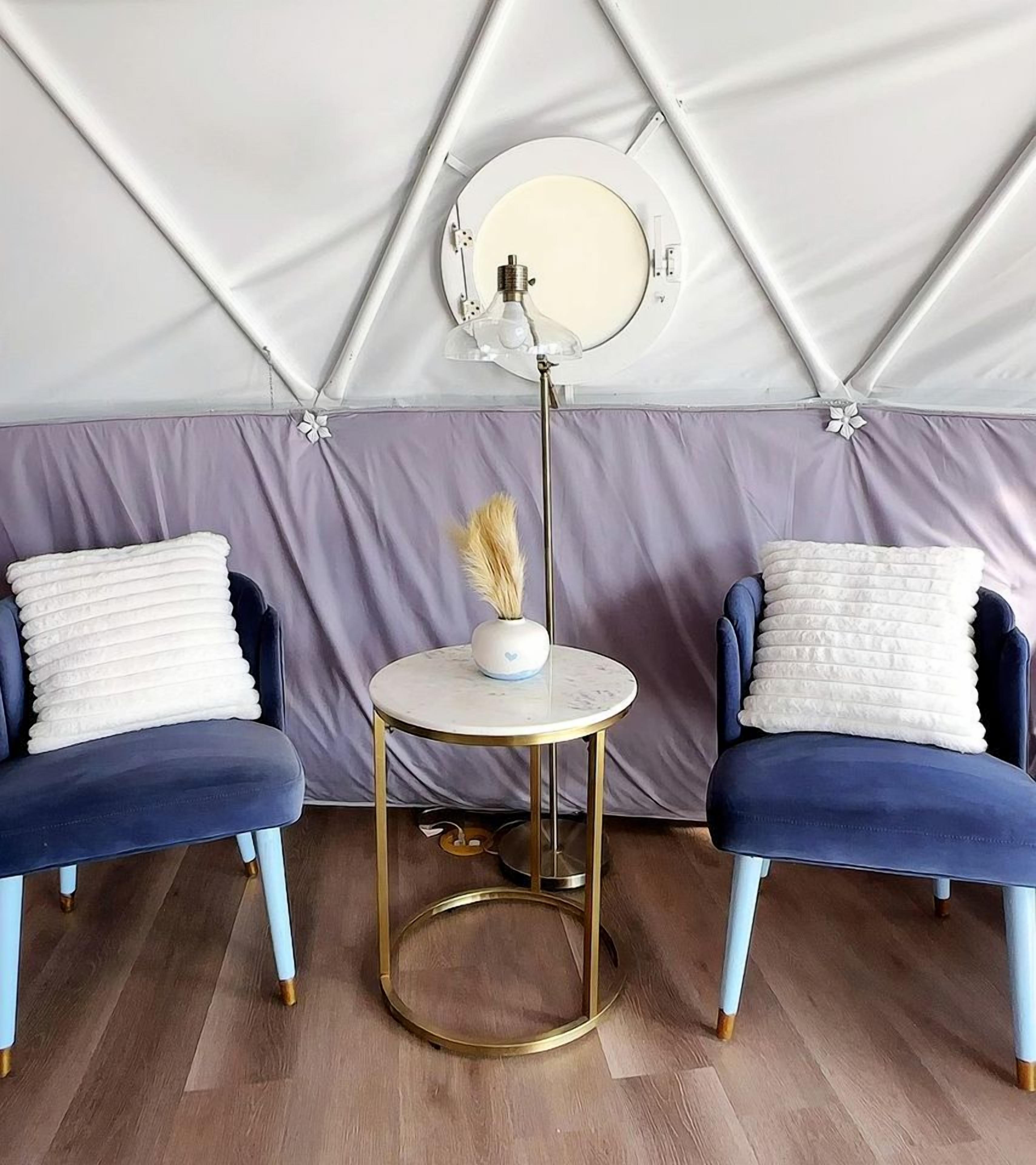 A tented room with two blue chairs, a round table, and a wall-mounted lamp.