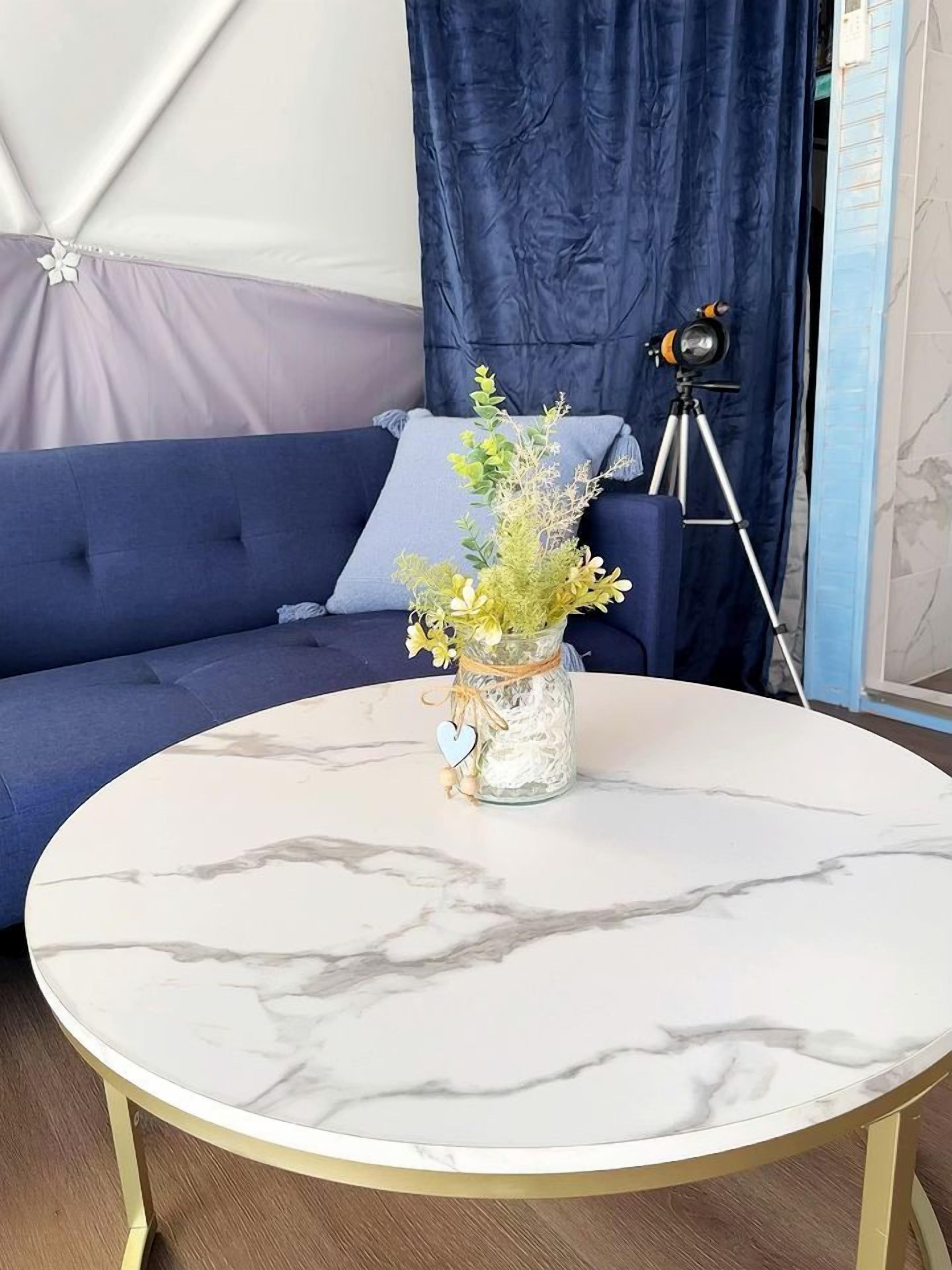 Une table ronde en marbre, sur laquelle se trouve un vase de fleurs, un trépied et un canapé bleu agrémenté d’un rideau, dans une pièce.