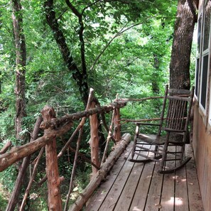 Um deck de madeira com uma cadeira de balanço, cercado por uma floresta densa.