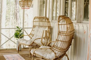 Two wicker chairs with white cushions on a wooden deck, a hanging light fixture, and a potted plant.