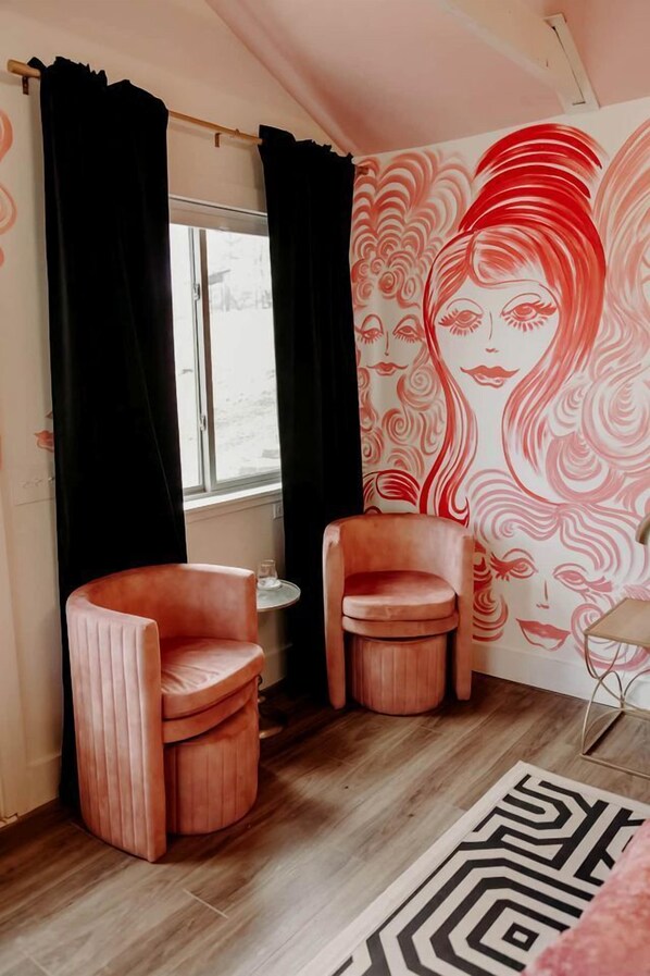 Una habitación con dos sillas rosadas, una mesita, un tapete estampado y un mural en la pared.