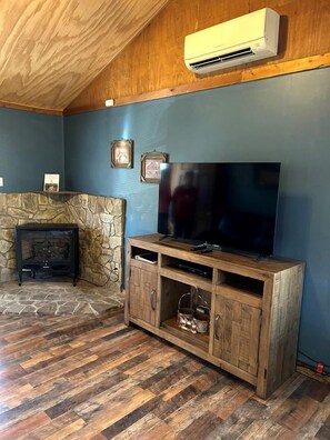 A living room with a stone fireplace, wooden entertainment unit, and an air conditioning unit mounted on the ceiling. - Pet-Friendly Cabin in North Carolina for Amazing Getaways in the Mountains (Spruce Pine)
