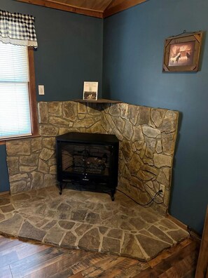 A stone fireplace with a black wood-burning stove, a framed picture on the wall, and a window with blinds. - Pet-Friendly Cabin in North Carolina for Amazing Getaways in the Mountains (Spruce Pine)