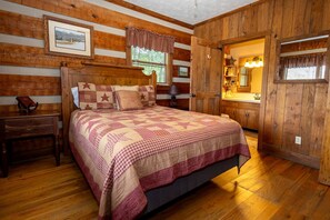 Une chambre à coucher aux murs lambrissés, avec un lit, une table de chevet, une lampe et une fenêtre agrémentée d’une lambrequin à carreaux.