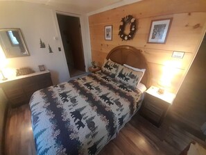 A bedroom with a wooden headboard, a bed with a patterned bedspread, a nightstand with a lamp, and a wall-mounted wreath.