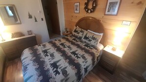 A bedroom with a wooden headboard, a bed with a patterned bedspread, a nightstand with a lamp, and a wall-mounted wreath. - Cozy Cabin Getaway in the Adirondack Mountains in Blue Mountain Lake, New York (Indian Lake)