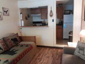 Una sala de estar acogedora con un sofá y un sillón, una cocina visible a través de un vano abierto, y una mesita con una lámpara.