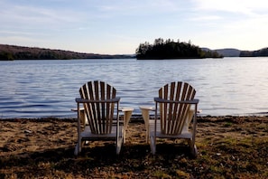 Two Adirondack chairs facing a lake with a distant island and forested hills.