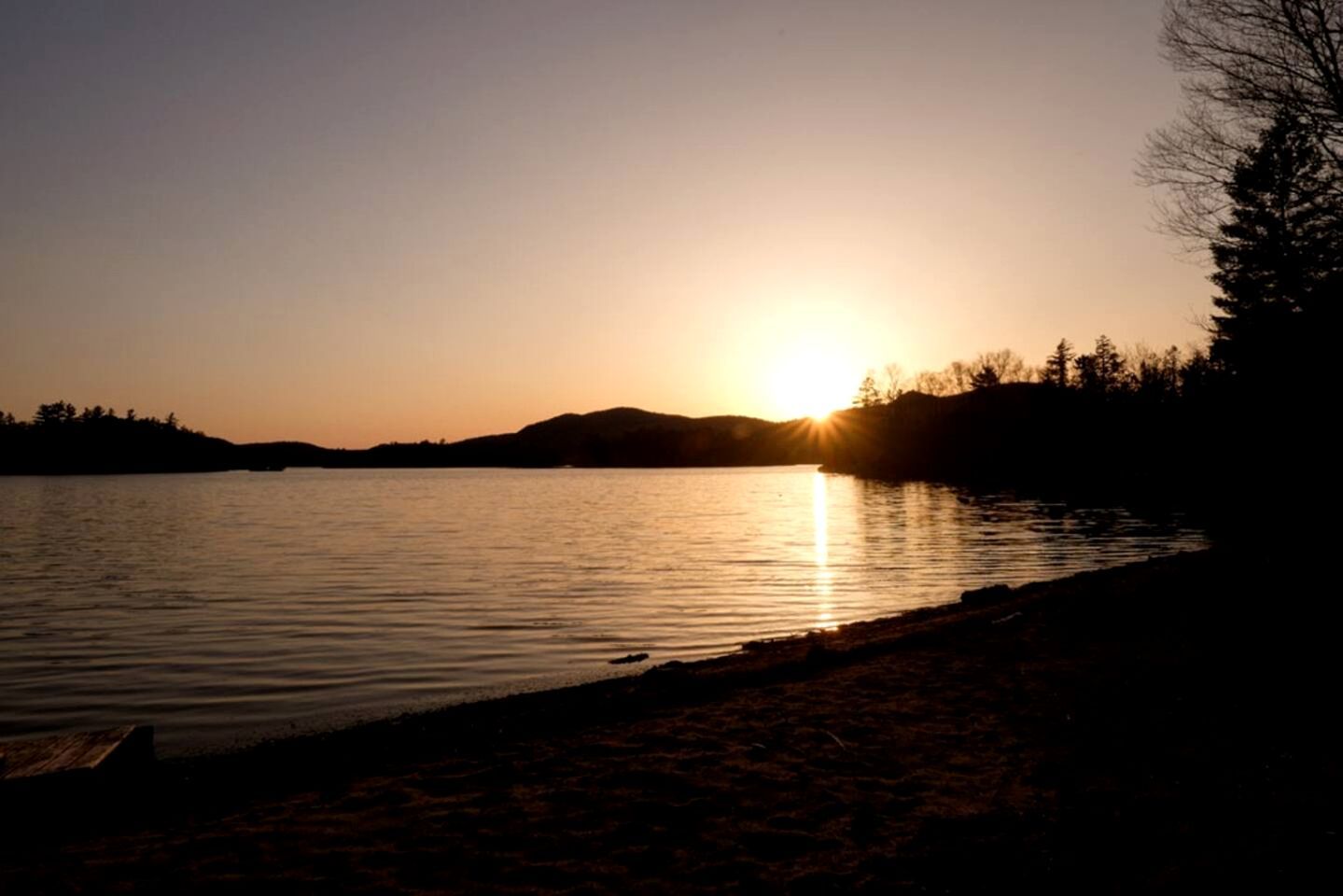 A sunset over a calm lake with a silhouette of trees and a distant hill.