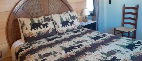 Una cama de madera con colcha adornada con motivos de bosque y osos, una mesita de noche con lámpara, y una silla en una habitación con paredes de madera y una ventana.
