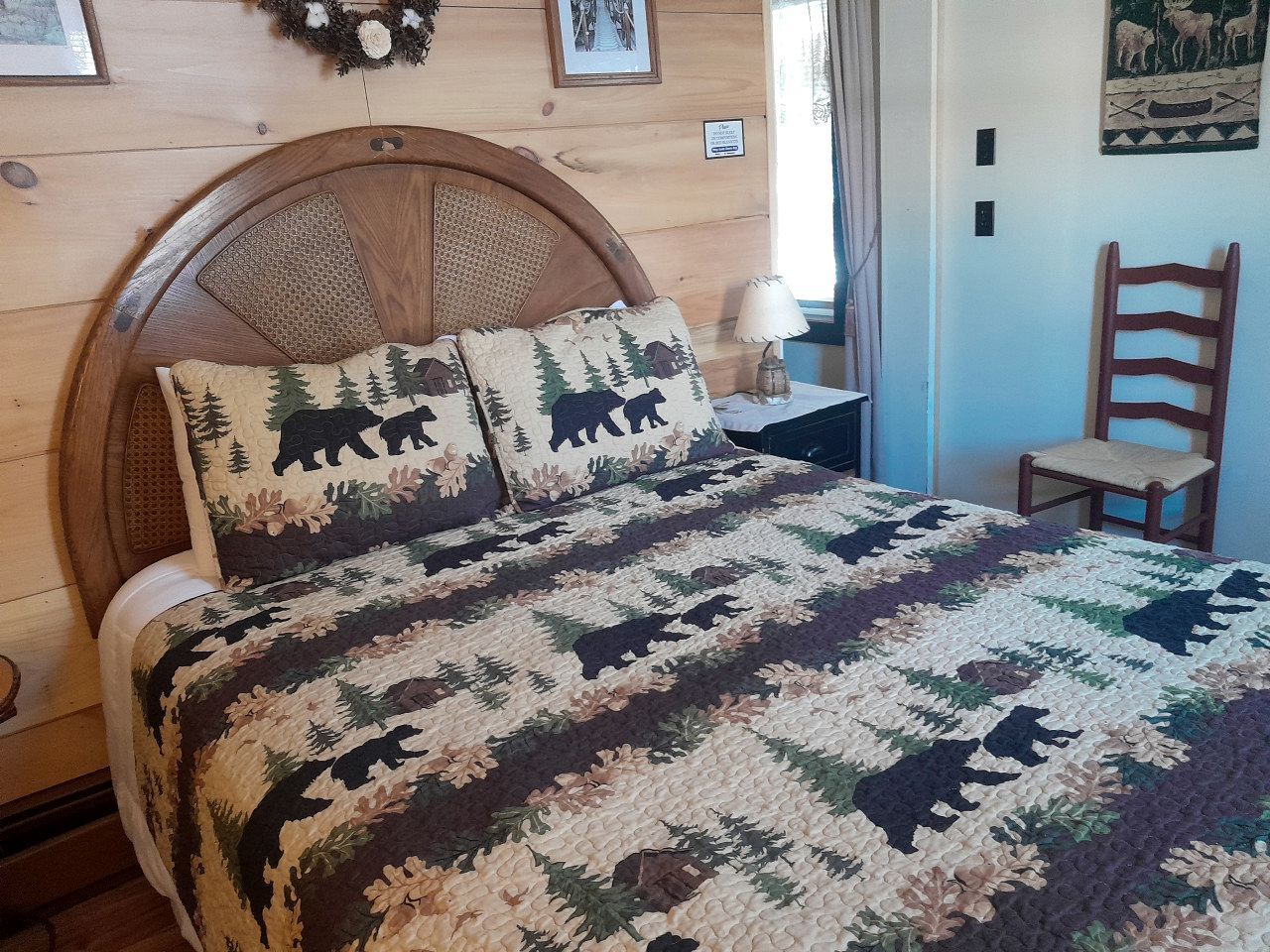 A wooden bed with a quilt featuring forest and bear patterns, a bedside table with a lamp, and a chair in a room with wooden walls and a window.