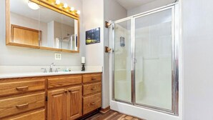 A bathroom with wooden cabinets, a white countertop, a large mirror with lights, and a glass shower enclosure.