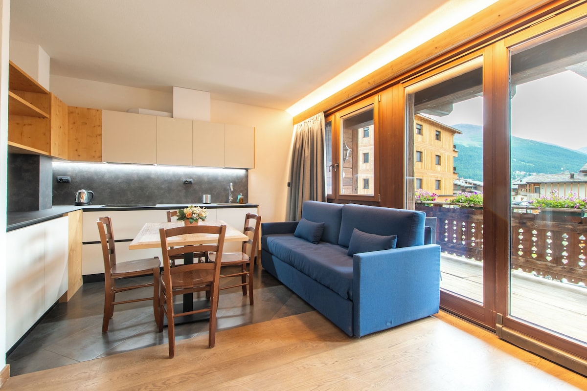 A modern kitchen with wooden cabinets, a dining table with chairs, and a blue sofa by a large window with a view of buildings and mountains.